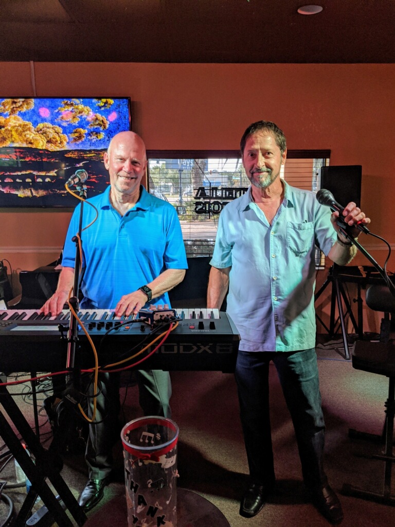 A balding guy in a blue shirt playing the piano while a bearded man in a light blue polo holds the mic to sing