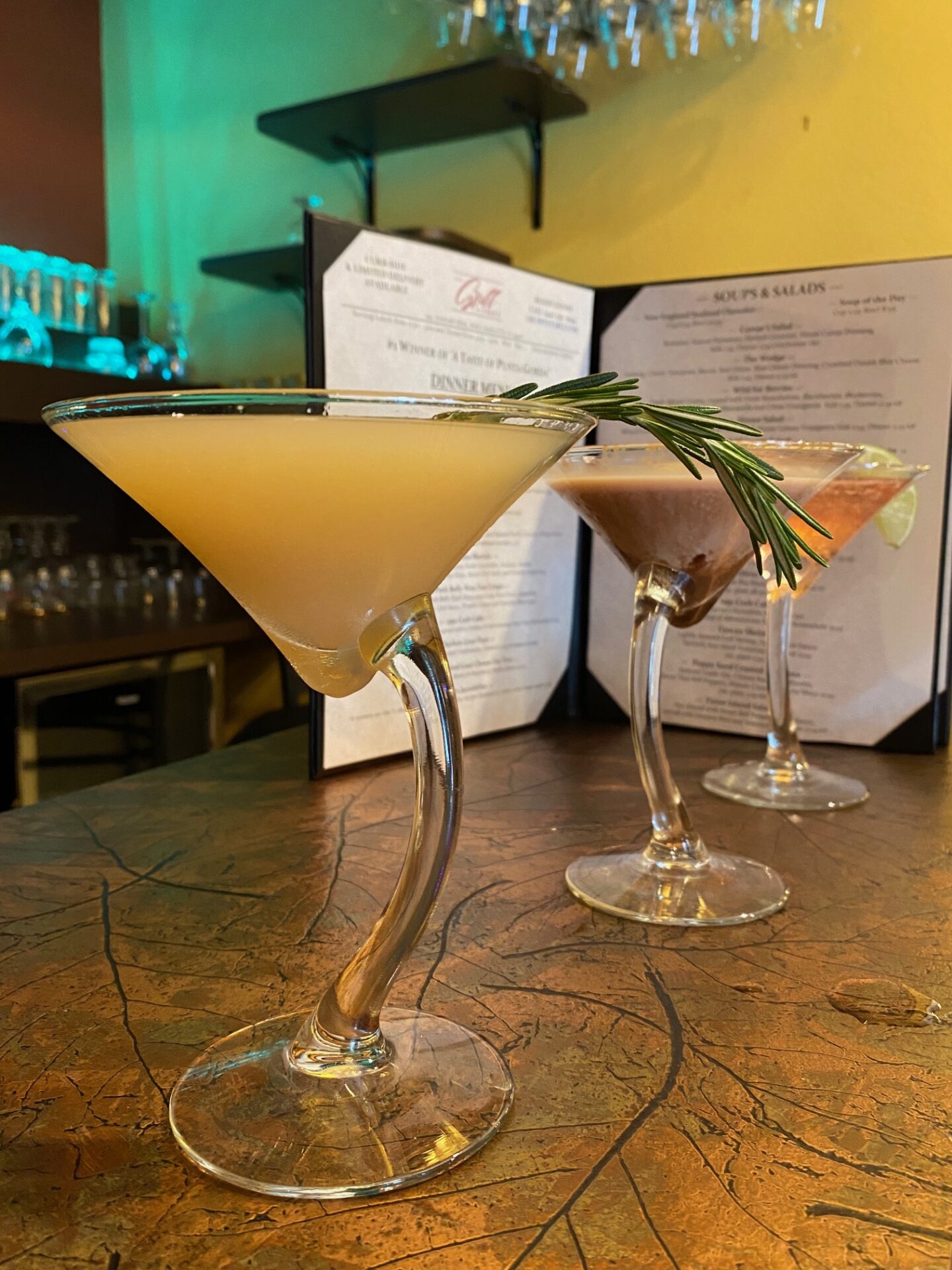 Three cocktail drinks in martini glasses with a curved handled placed on top of a bar table near an opened menu