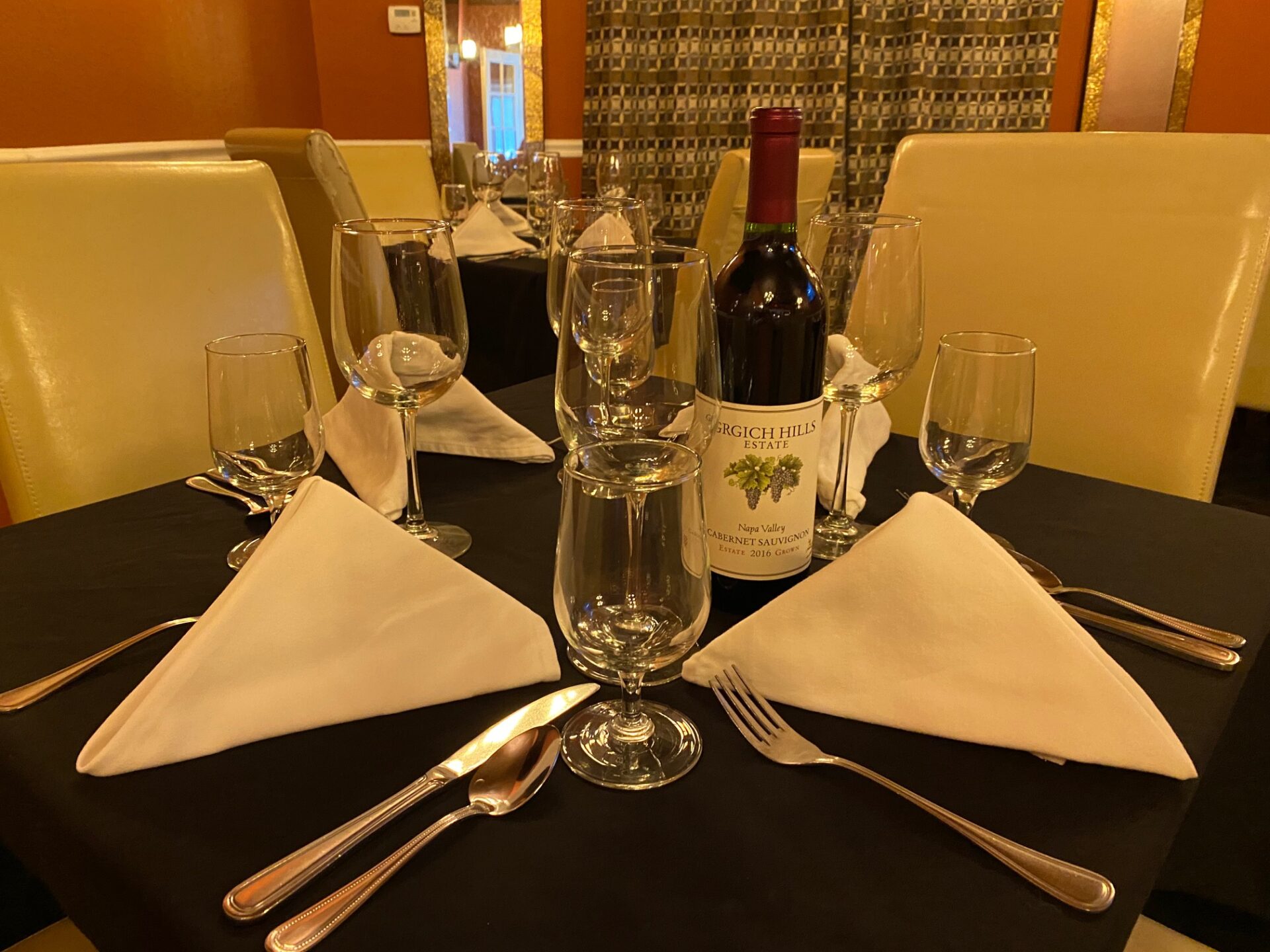 Table covered in a black cloth and set up with napkins, utensils, wine glasses, and a wine bottle